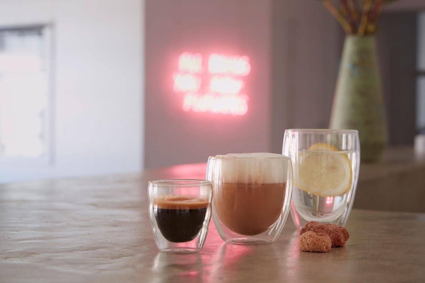 Three espresso shot glasses of varying sizes in front of a pink neon sign